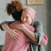 Foto de duas mulheres se abraçando, uma está com lenço na cabeça, como costumam usar as pacientes de câncer. O cuidado é essencial.
