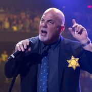 Foto do cantor e compositor Billy Joel, num palco, com microfone na mão e dedo em riste, com a estrela de Davi no peito, exemplo de homem valente