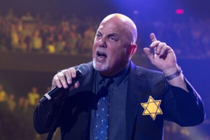 Foto do cantor e compositor Billy Joel, num palco, com microfone na mão e dedo em riste, com a estrela de Davi no peito, exemplo de homem valente