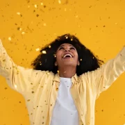 Foto de uma mulher negra jovem, alegre, recebendo uma chuva de confete. O recado é: celebre as conquistas.