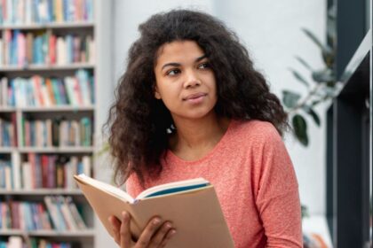 Foto mostra mulher negra clara, com cabelos compridos encaracolados, numa biblioteca, tem um livro nas mãos, mas olha pela janela, como se pensasse sobre a fúria do mundo