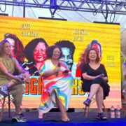Foto tem 3 mulheres sentadas em banquetas altas num palco com fundo amarelo, e um homem à direita, que faz a mediação de um debate sobre o futuro.