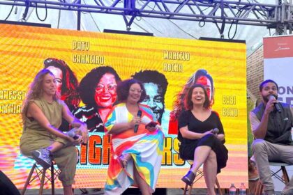Foto tem 3 mulheres sentadas em banquetas altas num palco com fundo amarelo, e um homem à direita, que faz a mediação de um debate sobre o futuro.
