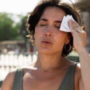 Imagem de uma mulher, na rua, suspirando de calor, passando um lenço na testa, são sintomas da menopausa