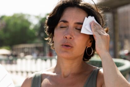 Imagem de uma mulher, na rua, suspirando de calor, passando um lenço na testa, são sintomas da menopausa