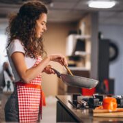Foto de mulher jovem, cabelos compridos e meio crespos, com avental vermelho, está no fogão fazendo comida.