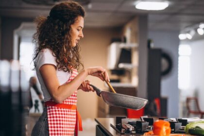 Foto de mulher jovem, cabelos compridos e meio crespos, com avental vermelho, está no fogão fazendo comida.