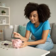 Foto de uma mulher sentada no sofá, colocando moedas no seu cofre de porquinho rosa. Organização Financeira é isso.