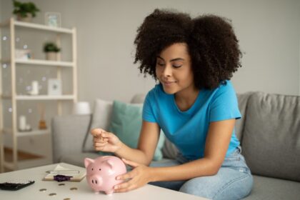 Foto de uma mulher sentada no sofá, colocando moedas no seu cofre de porquinho rosa. Organização Financeira é isso.