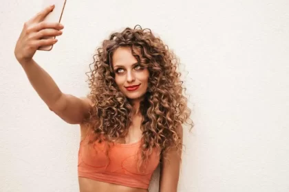 Foto de uma mulher jovem, bonita, cabelos longo, claro e encaracolados, faz uma selfie com o fone celular. É a típica perfeitinha do Instagram.
