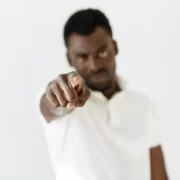 Foto num cenário branco, com um homem negro, de branco, apontando o dedo na direção da câmera, como se apontasse para um alguém preconceituoso