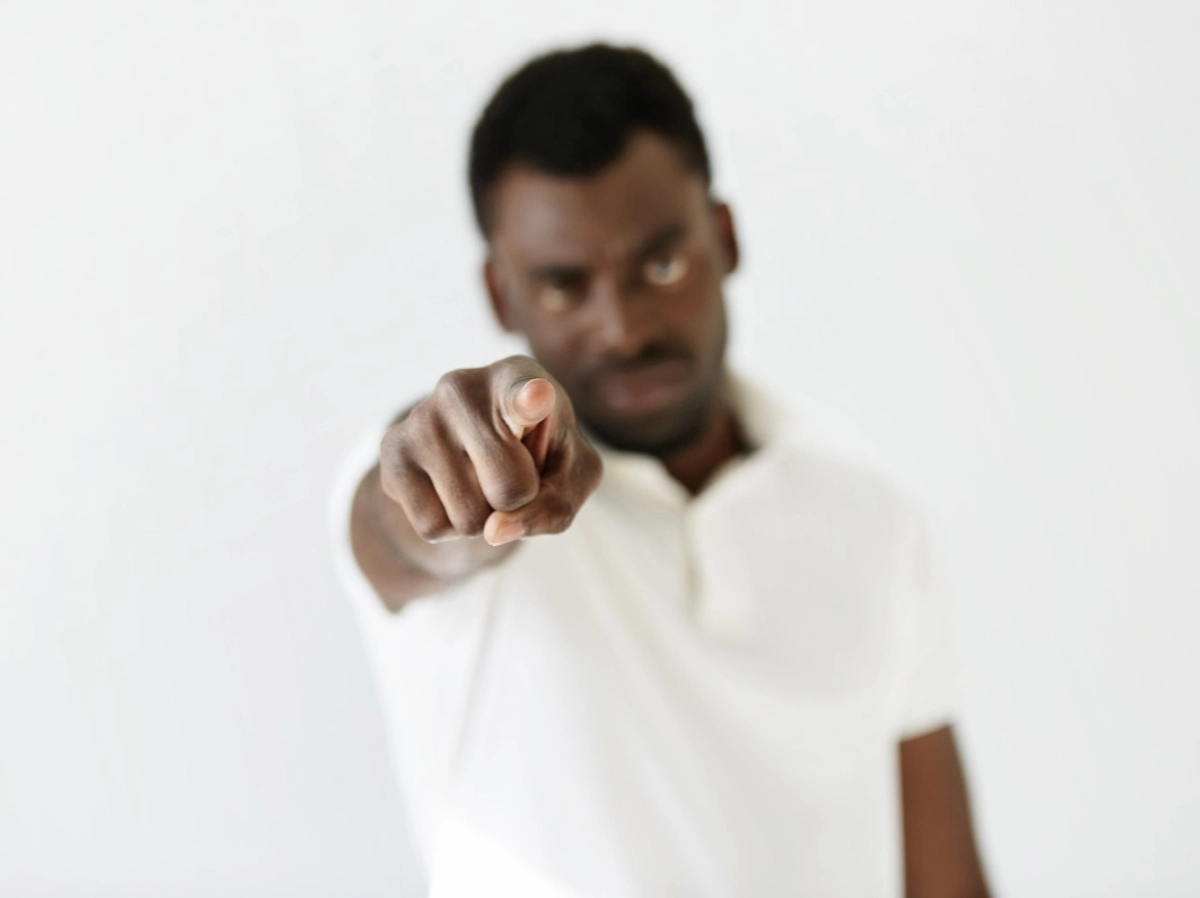 Foto num cenário branco, com um homem negro, de branco, apontando o dedo na direção da câmera, como se apontasse para um alguém preconceituoso