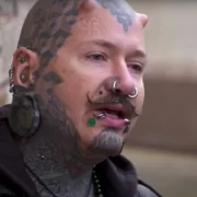 Foto de um homem de cerca de 35 anos com o rosto cheio de tatuagens, piercings e implantes. É um integrante da biblioteca humana.