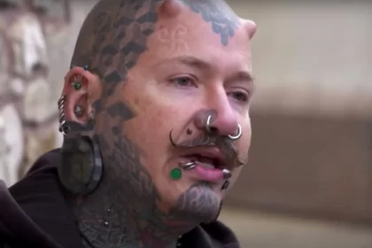 Foto de um homem de cerca de 35 anos com o rosto cheio de tatuagens, piercings e implantes. É um integrante da biblioteca humana.