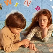 Foto de duas crianças, um menino e uma menina, deitados de bruços no chão, brincando. O menino sob o olhar da menina.