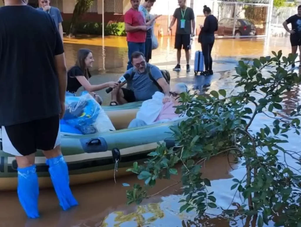 Foto de uma senhora sendo salva num bote (ou num berço) por voluntários, na enchente de porto alegre