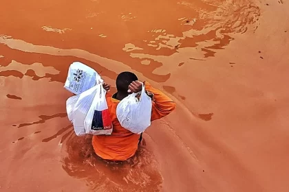 Foto de um homem com uma água barrenta até a cintura, carrega sacolas plásticas erguidas acima do ombro. Exemplo de refugiados do clima da enchente do RS