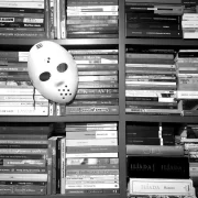 Foto em preto e branco de uma estante cheia de livros colocados horizontalmente, com uma máscara de terror pendurada no meio