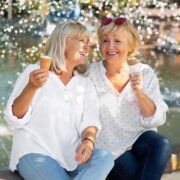 Duas mulheres maduras tomam sorvete e riem, sentadas na beira de uma fonte de água, pessoas