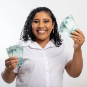 Foto de mulher negra clara, sorrindo, mostrando notas de 100 reais nas duas mãos. É um apelo ao concumo consciente