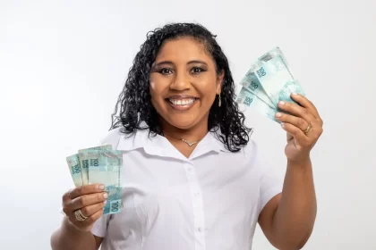 Foto de mulher negra clara, sorrindo, mostrando notas de 100 reais nas duas mãos. É um apelo ao concumo consciente