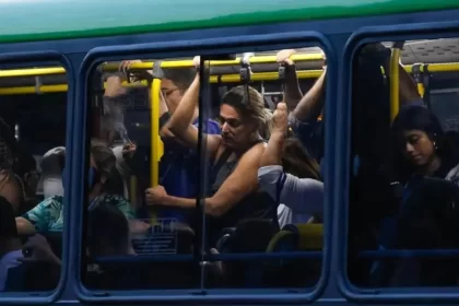 Foto da janela de um ônibus cheio em deslocamento, com pessoas apertadas. Corpo espremido.