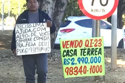 Homem de aparência humilde carrega um cartaz pedindo ajuda por pix e ao lado um cartaz anunciando uma mansão à venda.