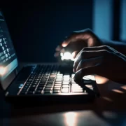 Foto de um ambiente escuro. As mãos de um homem estão sobre o teclado de um lap top. Deputados precisam votar lei das fake news