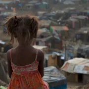 Imagem gerada por IA mostra menina parda de costas de uma área elevada, olhando para um acampamento de refugiados. É o cansaço civilizacional.