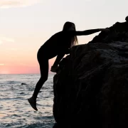 Foto num fim de tarde, mulher escalando uma pedra grande na beira do mar. Há quem busque prazer no risco