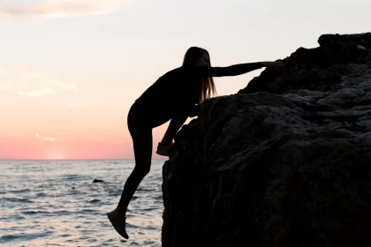 Foto num fim de tarde, mulher escalando uma pedra grande na beira do mar. Há quem busque prazer no risco