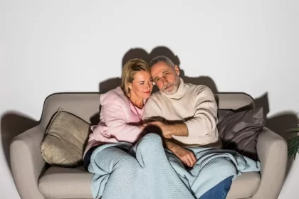 Foto de um casal baraçado, sentado num sofá, com cobertor cobrindo as pernas. É o amor.