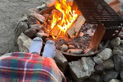 Foto mostra as pernas de uma pessoas com cobertor e os pés com meia próximos a uma fogueira. Montanhas ao fundo.