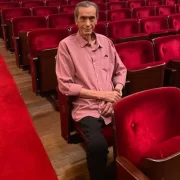 Foto do compositor Verocai, sentado numa cadeira de uma plateia de um teatro