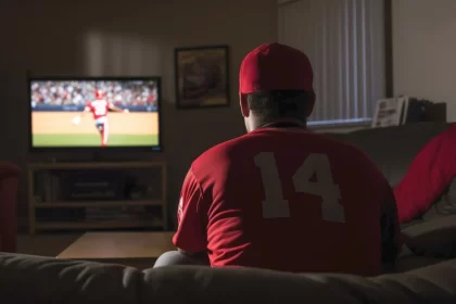 Foto de um homem de costas, sentado no sofá em frente a uma TV, onde passa um jogo. Aposta nas BETs