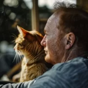 Foto de um homem sentado, em frente a janela com sol, e um gato no colo