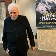 foto do cineasta Alejandro Jodorowsky, em frete a um cartaz de uma de suas retrospectivas