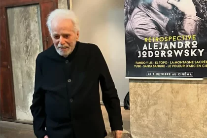 foto do cineasta Alejandro Jodorowsky, em frete a um cartaz de uma de suas retrospectivas