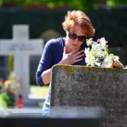 Uma mulher em frente a um túmulo, com flores na mão e expressão de tristeza. Dia de Finados