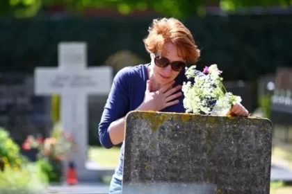 Uma mulher em frente a um túmulo, com flores na mão e expressão de tristeza. Dia de Finados