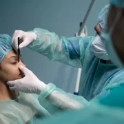 Foto mostra um ambiente cirúrgico, um médico está com mas mãos no rosto de uma paciente