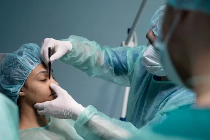 Foto mostra um ambiente cirúrgico, um médico está com mas mãos no rosto de uma paciente