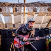 Foto do cantor e compósito Charles Master, em cima de um palco, de óculos escuros e boné, segurando uma guitarra