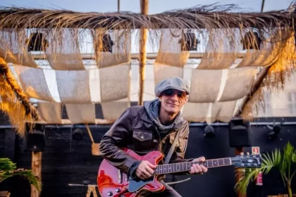 Foto do cantor e compósito Charles Master, em cima de um palco, de óculos escuros e boné, segurando uma guitarra