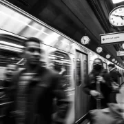 Foto p/b de uma estação de metro, trem passa rápido, pessoas com pressa, vários relógios. O autor discute o tempo e a pressa