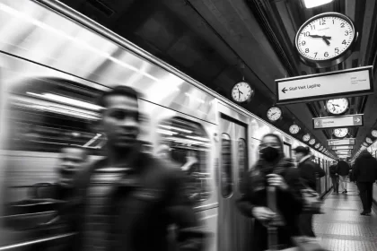 Foto p/b de uma estação de metro, trem passa rápido, pessoas com pressa, vários relógios. O autor discute o tempo e a pressa