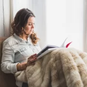 Mulher sentada, encostada numa parede, com um cobertor sobre as pernas, lendo um livro