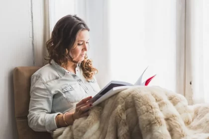 Mulher sentada, encostada numa parede, com um cobertor sobre as pernas, lendo um livro