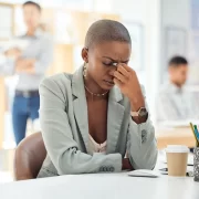 Mulher negra, com cabeça raspada, senata numa mesa com a mão nos olhos. Autora fala de assédio no trabalho