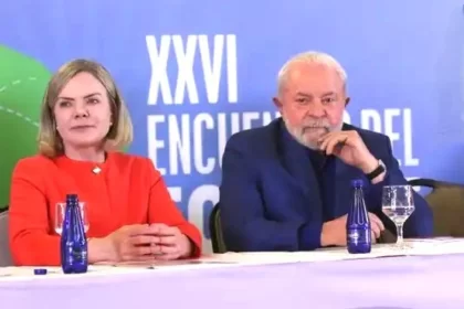 Foto de Lula e Gleisi Hoffmann sentados numa bancada de um evento